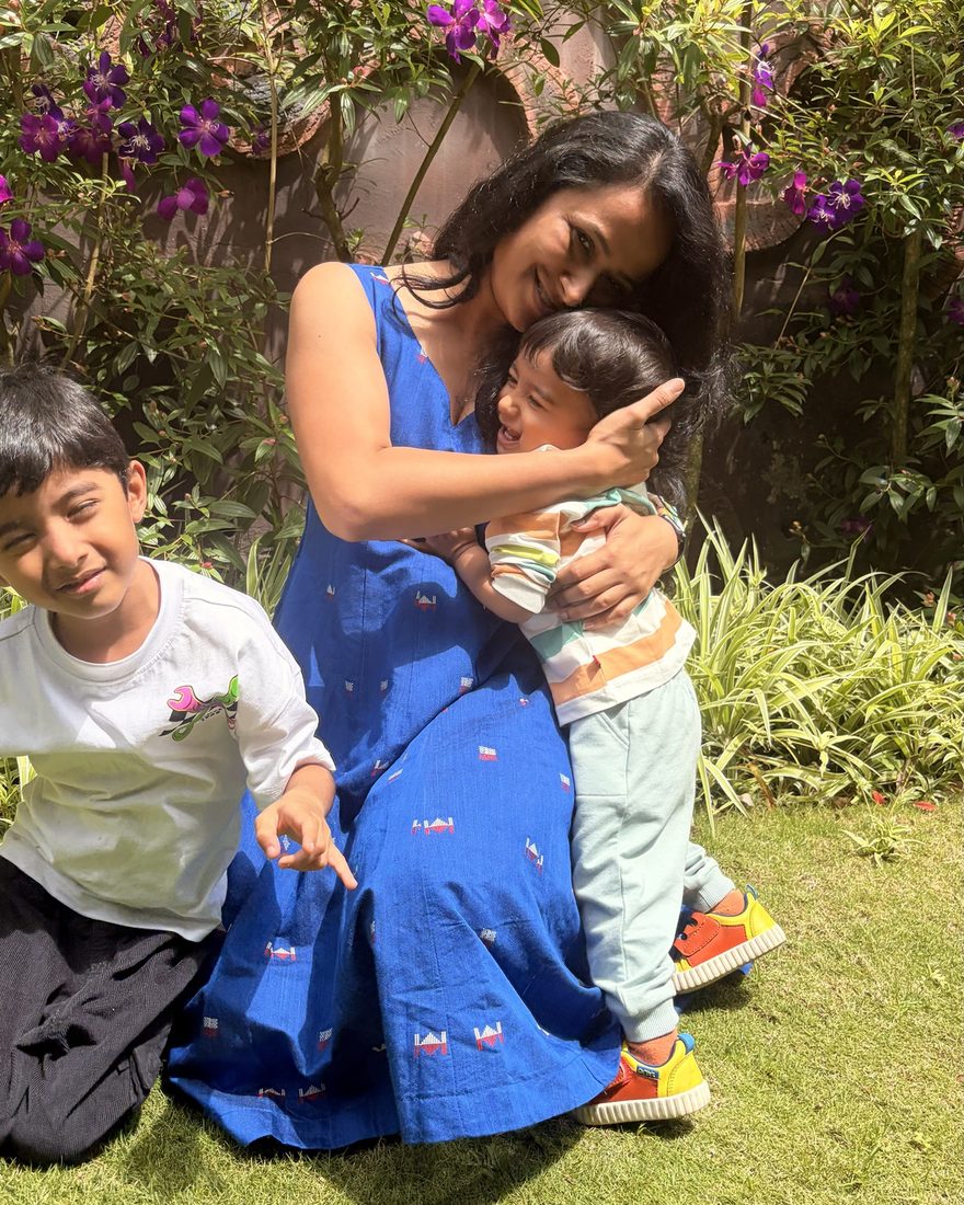 Manali hugging her younger son in a garden while her older son grins at the camera